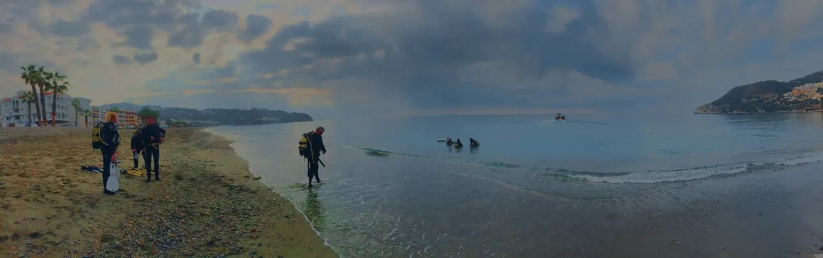 Panorámica de la bahía de La Herradura al amanecer: tres buceadores con la botella puesta esperan en la orilla mientras otro entra al agua, con un grupo ya dentro y la neumática al fondo.