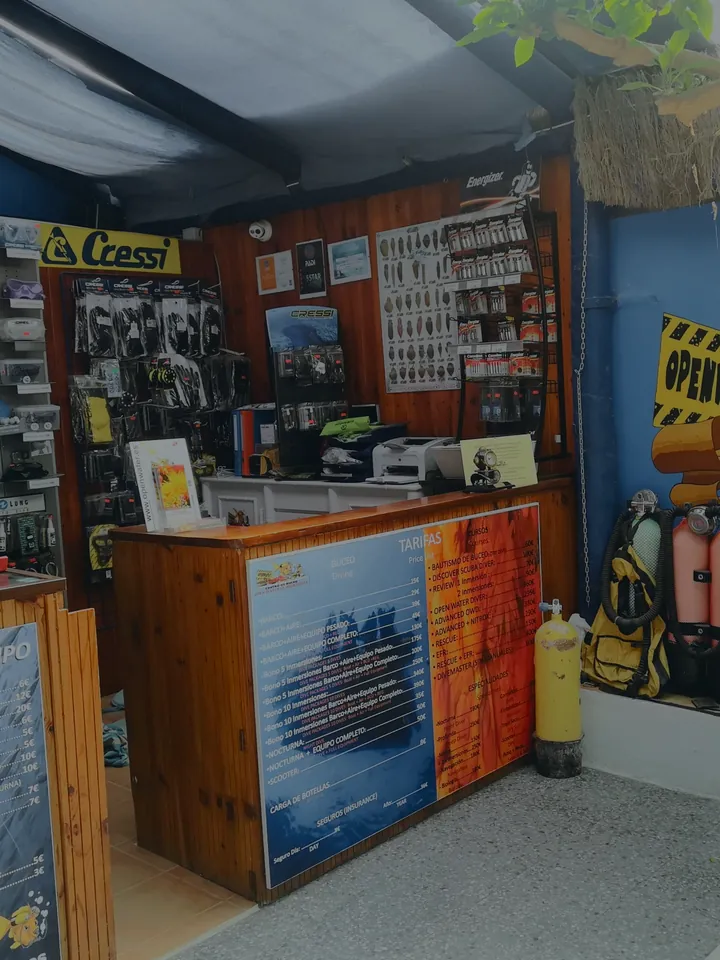 La tienda del centro por dentro: el mostrador de alquiler de equipo, la pared de reguladores y máscaras Cressi y botellas de colores bajo un mural con un pez.