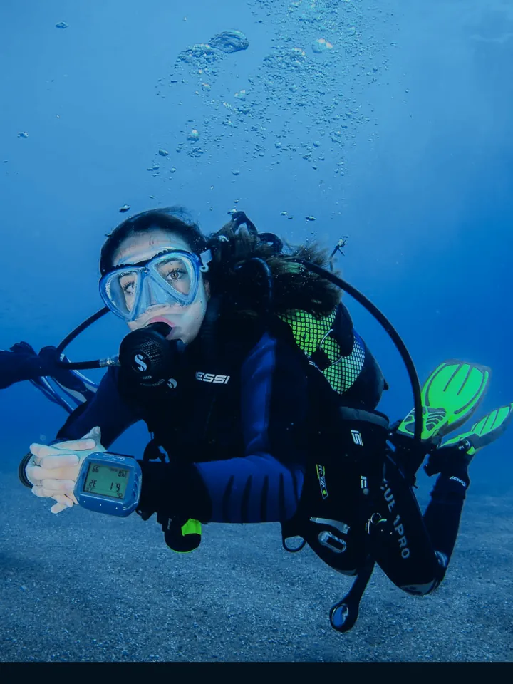 Una buceadora en el agua azul, con máscara y equipo Cressi, mira el ordenador de buceo de su muñeca.