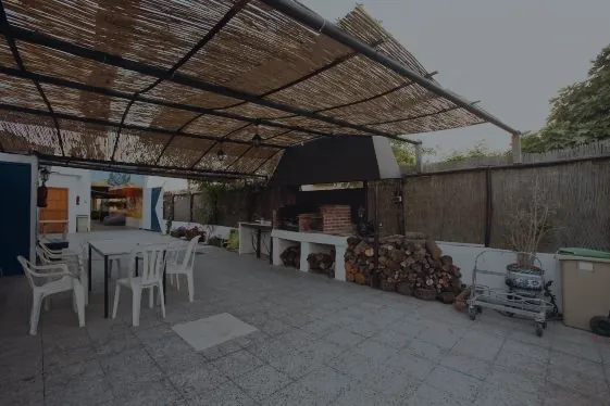 Terraza del centro bajo un techo de cañizo, con mesas y sillas de plástico y una barbacoa de obra con leña apilada al fondo.