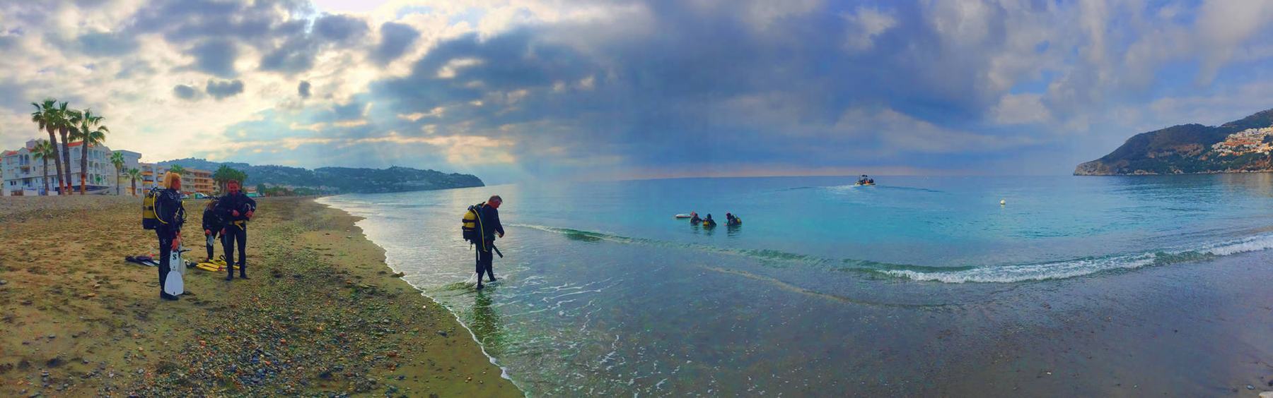 Panorámica de la bahía de La Herradura al amanecer: tres buceadores con la botella puesta esperan en la orilla mientras otro entra al agua, con un grupo ya dentro y la neumática al fondo.