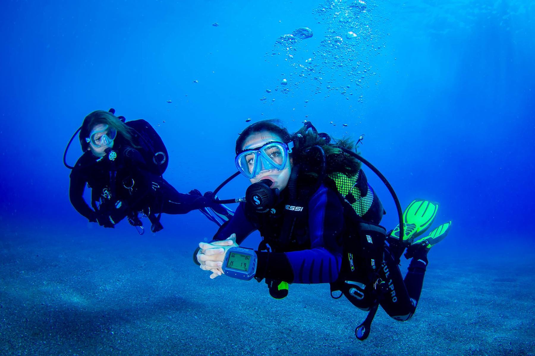 Dos buceadoras sobre el fondo de arena; la de delante mira el ordenador de buceo que lleva en la muñeca.