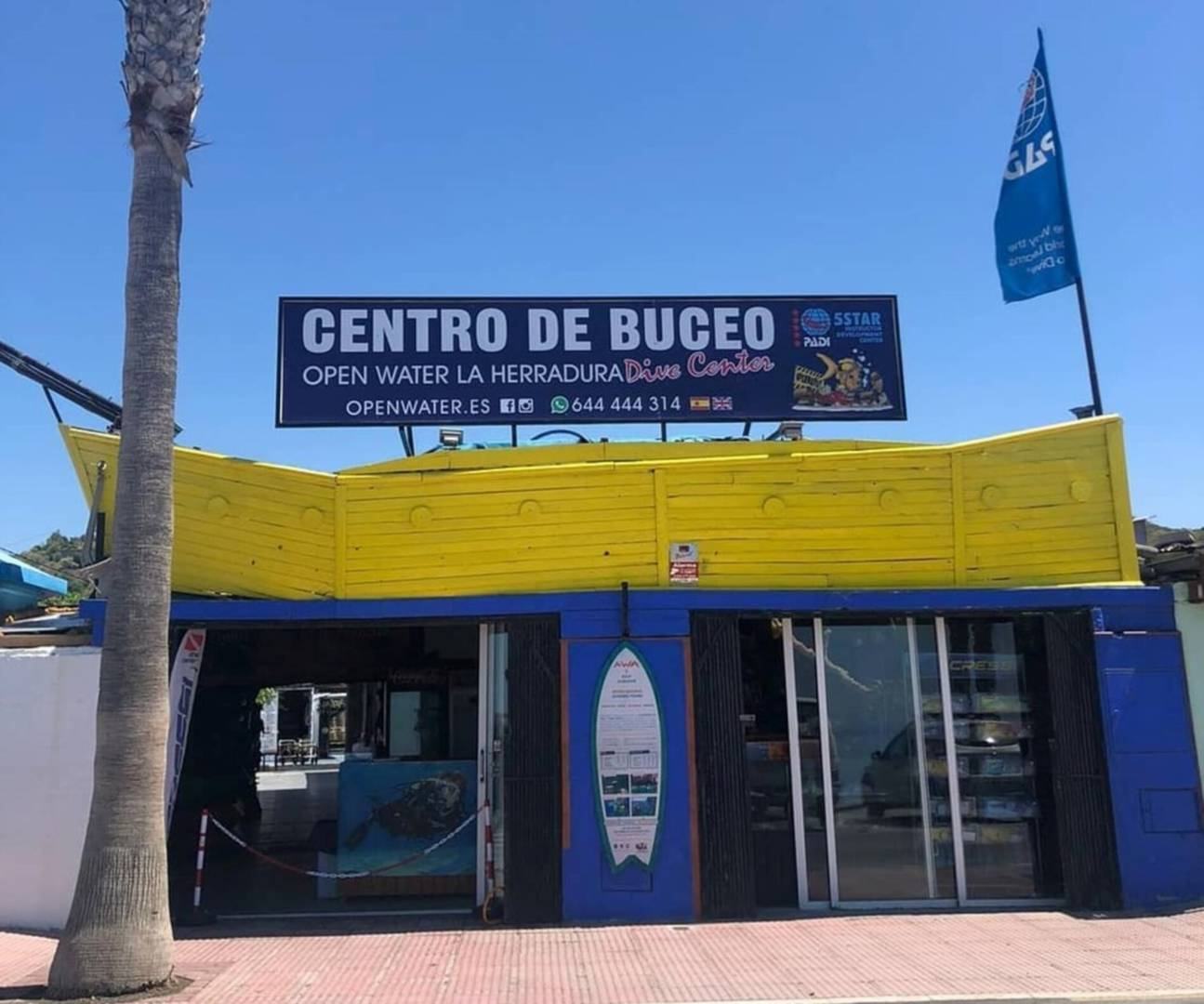 Fachada del centro de buceo en el paseo: el rótulo de Open Water La Herradura sobre un porche de madera amarilla, la tienda a pie de calle y una palmera delante.