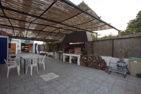 Terraza del centro bajo un techo de cañizo, con mesas y sillas de plástico y una barbacoa de obra con leña apilada al fondo.
